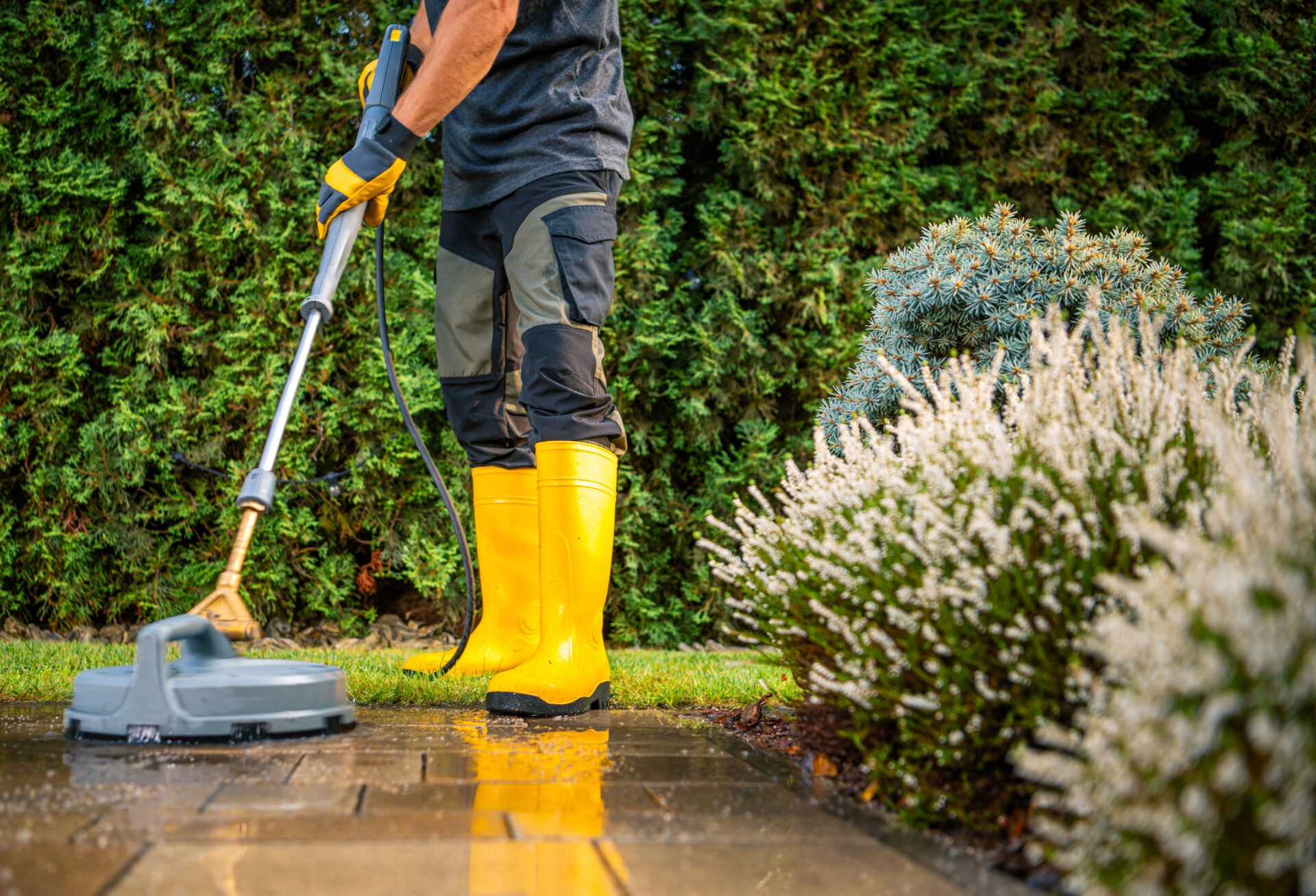 Person Uses a Pressure Washer to Clean Patio Stones While Wearing Yellow Boots and Standing on a Well-Maintained Lawn Near Blooming Bushes During a Sunny Afternoon